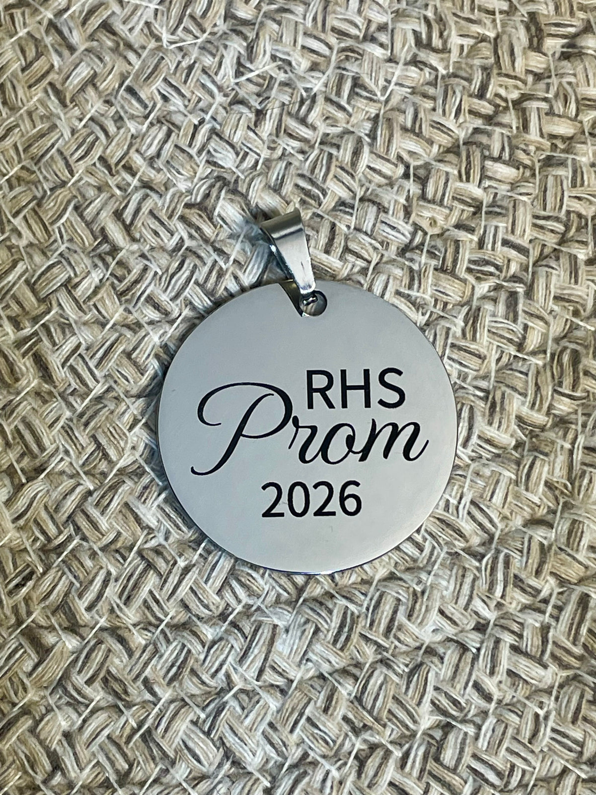 A round silver pendant engraved with 'RHS Prom 2026' in black lettering, resting on a textured beige woven mat background.