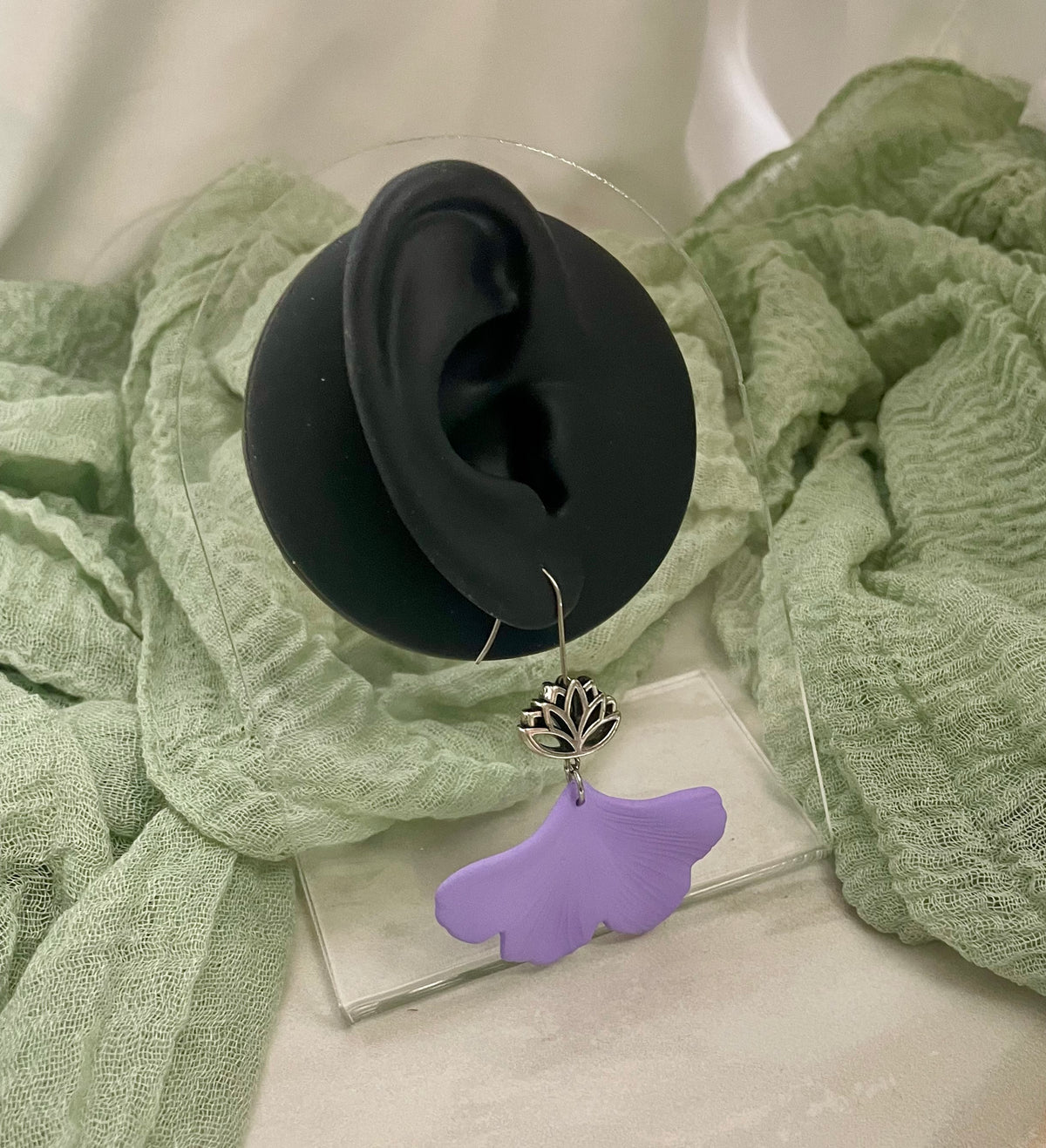 Purple gingko earring with lotus charm on a clear stand with a black ear model and green fabric in the background