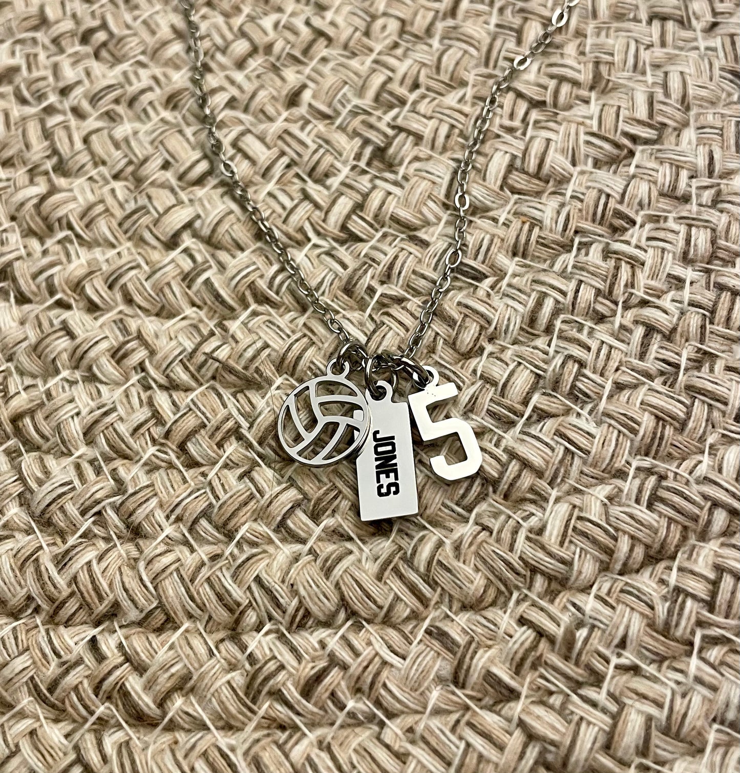 All silver necklace with volleyball charm, 'JONES' engraved tag and number 5 charm on a woven fabric background