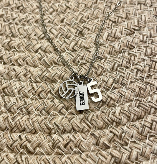 All silver necklace with volleyball charm, 'JONES' engraved tag and number 5 charm on a woven fabric background