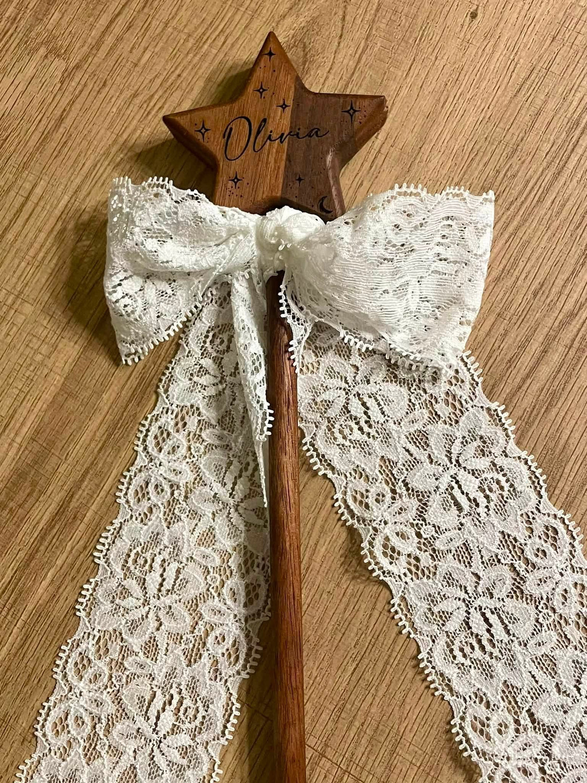 A wooden star wand with a carved name 'Olivia' and small star details, tied with a large white lace bow and long lace tails, placed on a light wood surface.