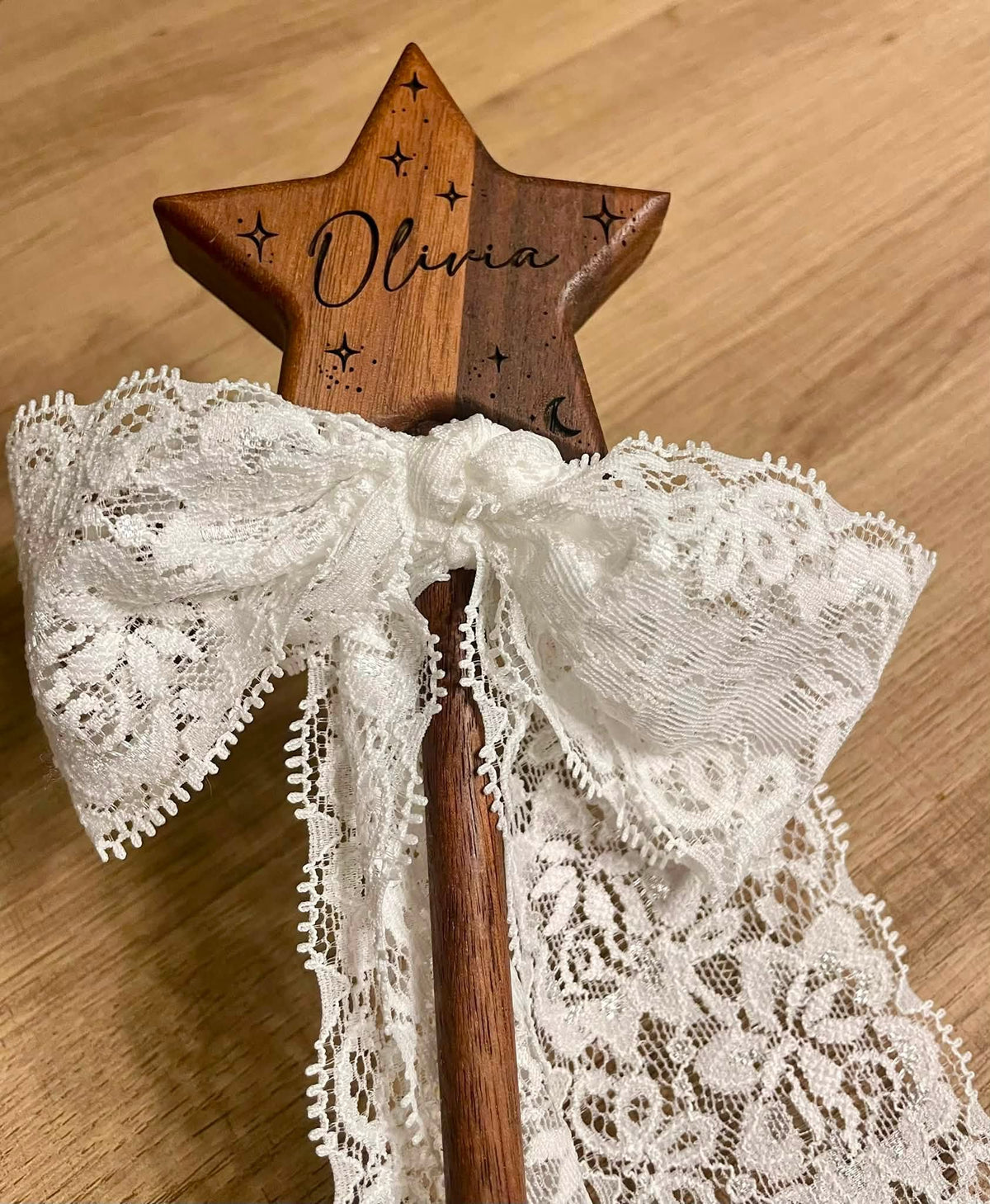 A wooden star wand topper with the name 'Olivia' engraved on the star, decorated with a large white lace bow and matching lace ribbon, shown on a wooden surface.