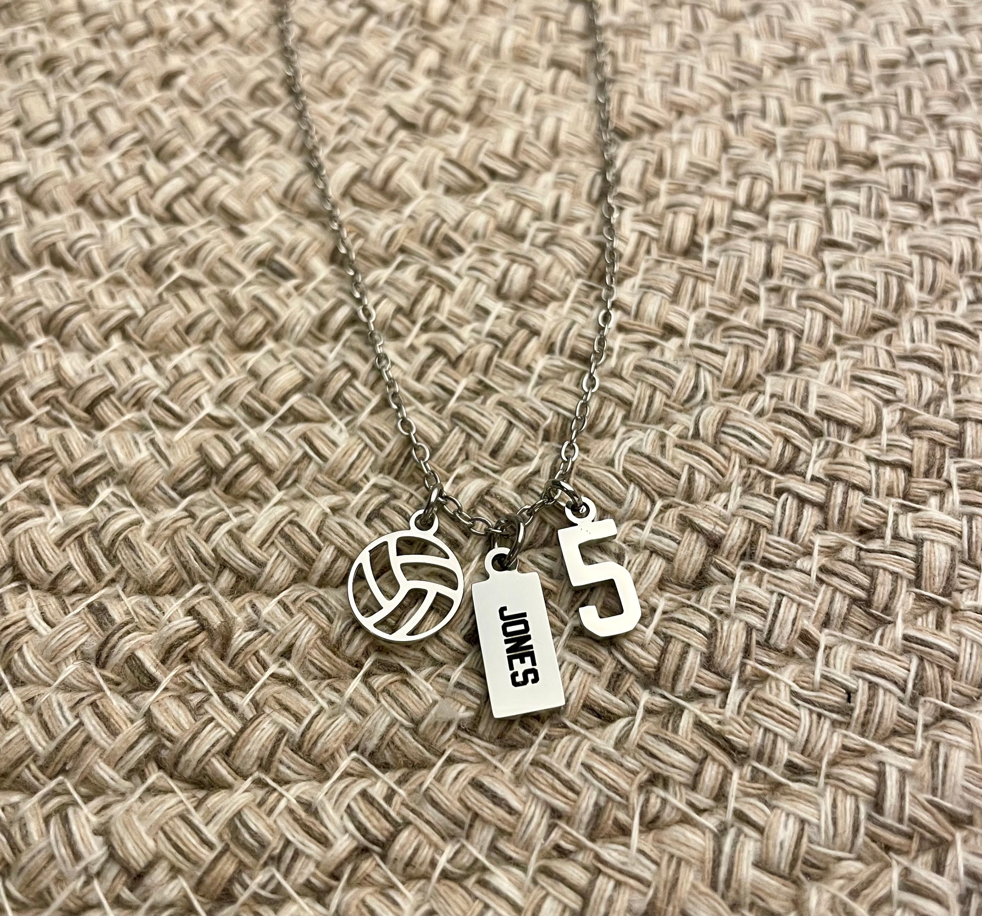 All silver necklace with volleyball charm, 'JONES' engraved tag and number 5 charm on a woven fabric background attached to not move on necklace