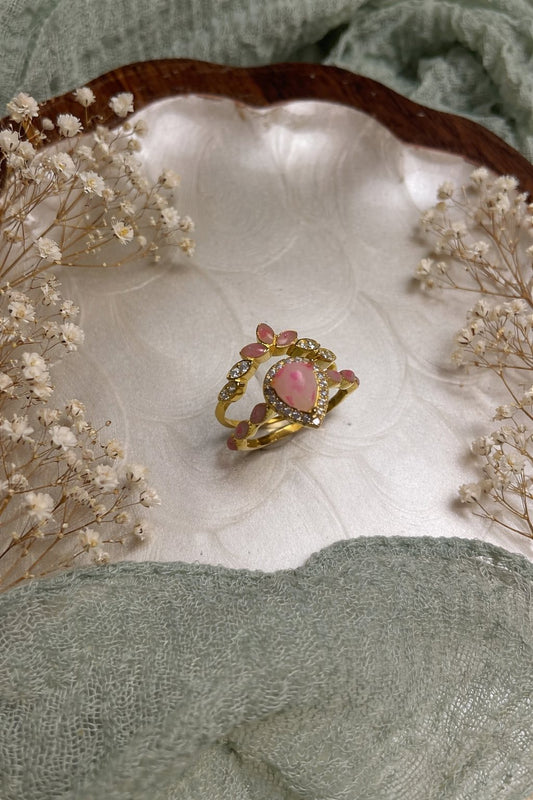 Gold ring with a pink flake & breast milk on a white shell surrounded by dried flowers, on a green fabric background.