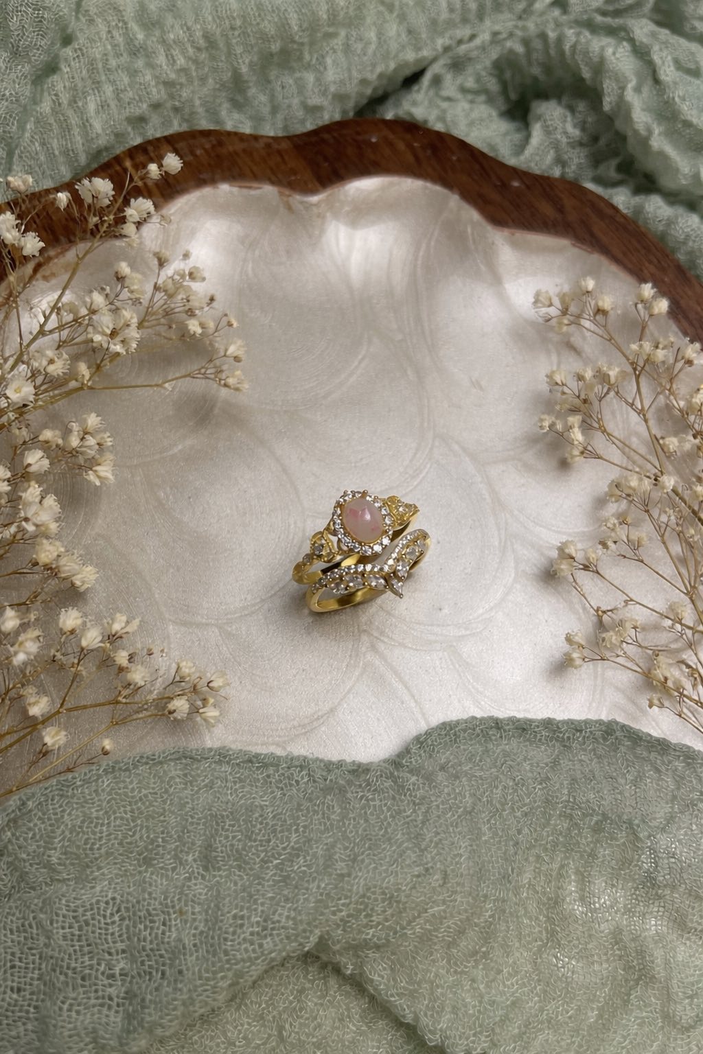 Gold ring with a pink flake & breast milk in a shell surrounded by dried flowers on a textured green fabric background