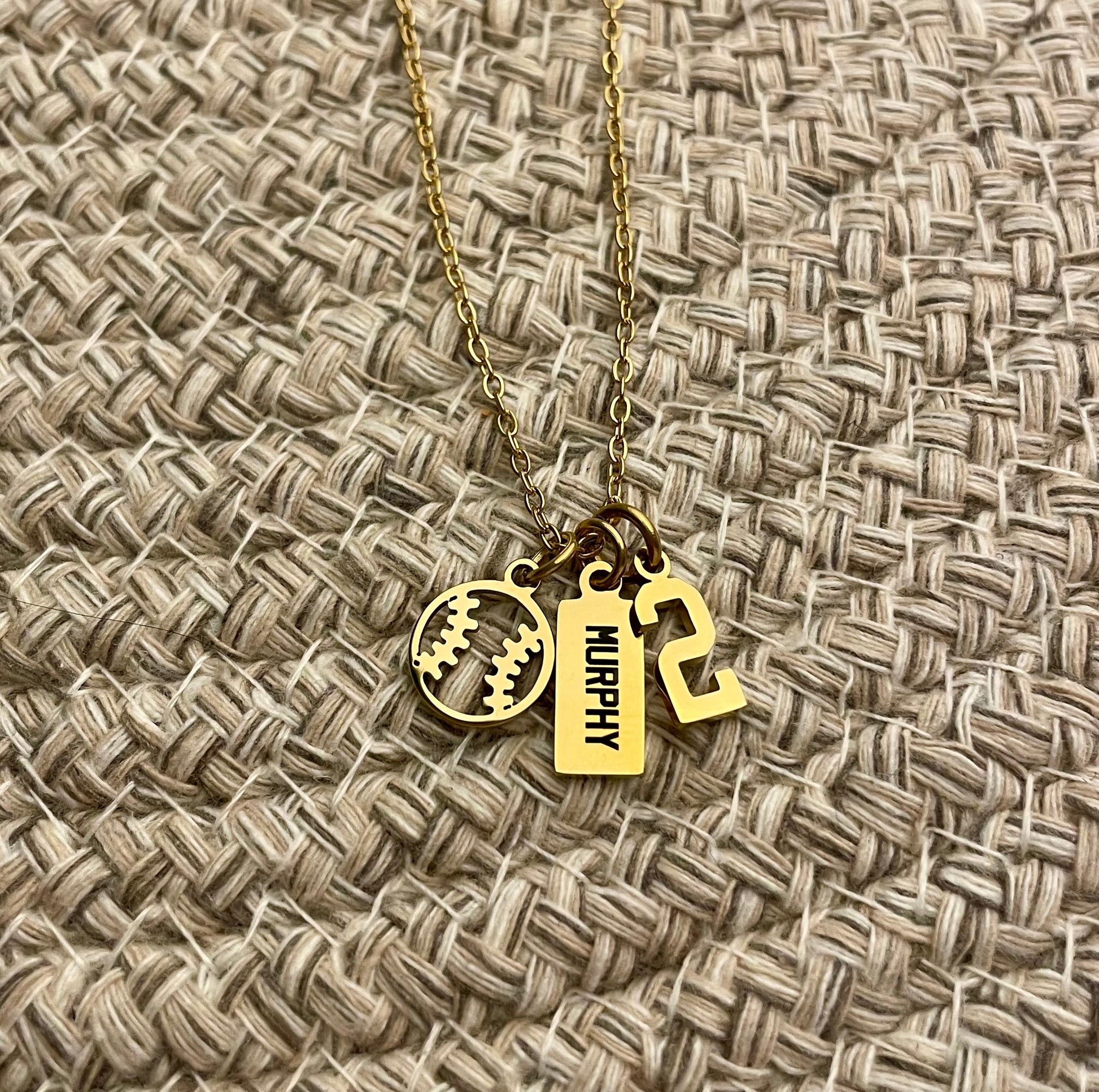 Gold necklace with softball charm, engraved 'MURPHY' tag and the number 2 charm on a textured beige background