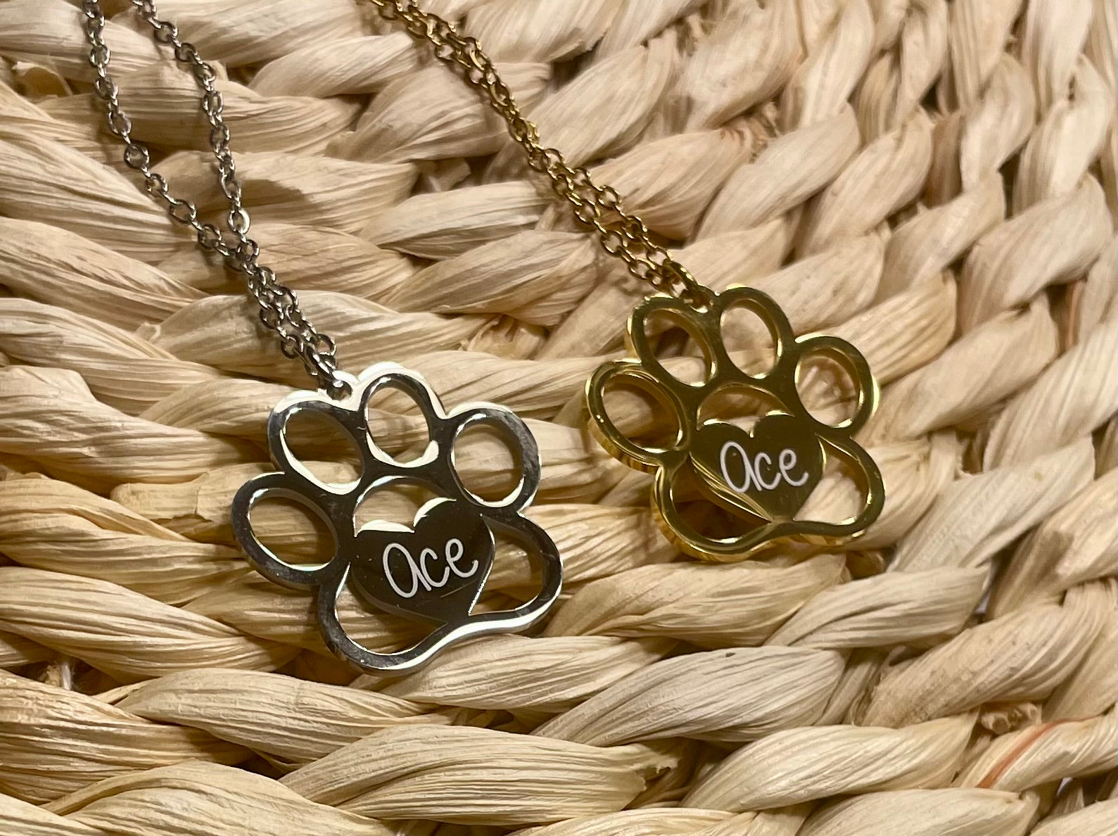 Two paw-shaped necklaces with 'Ace' engraved on them, one gold and one silver, on a woven surface.