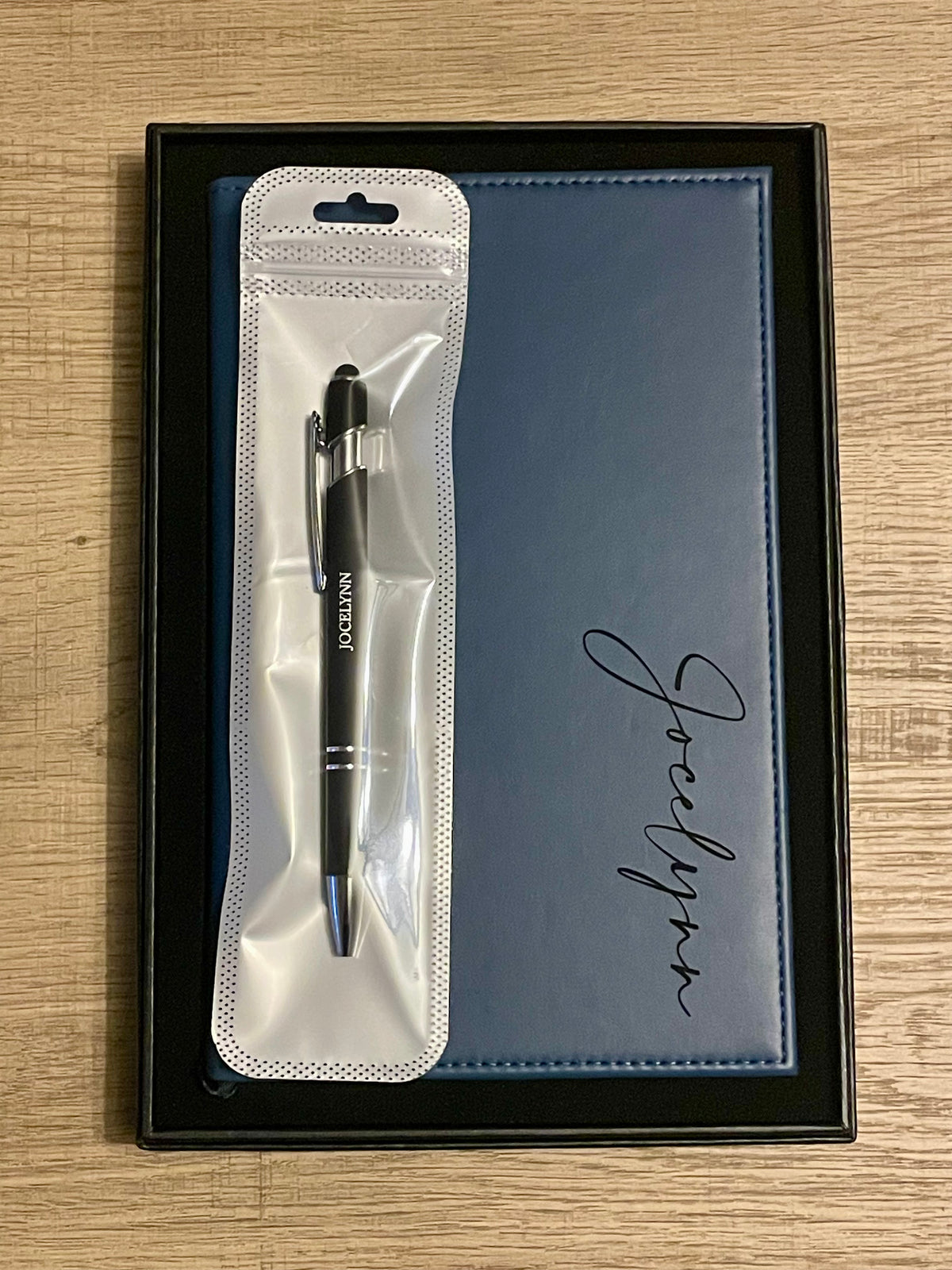 A black ballpoint pen in a clear plastic sleeve, presented next to a dark blue textured notebook both engraved, inside a black gift box on a light wood background.