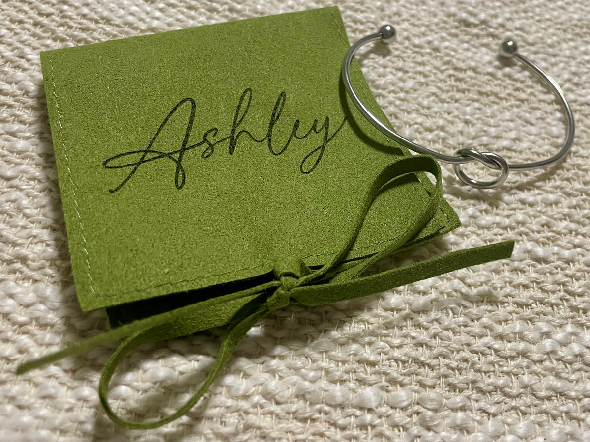 A silver metal cuff bracelet with ball-end caps, displayed next to a personalized olive green jewelry pouch that reads 'Ashley' in black script, tied with a matching ribbon on a textured cream woven fabric background.