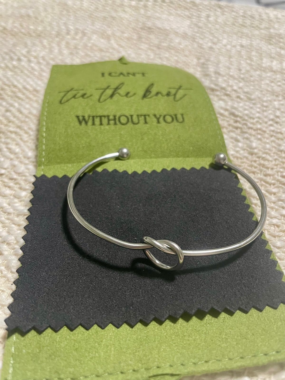 A silver-toned metal cuff bracelet with a knotted center detail, displayed on a black textured cleaning cloth over a green jewelry pouch. The engraved jewelry pouch reads 'I CAN'T tie the knot WITHOUT YOU'.