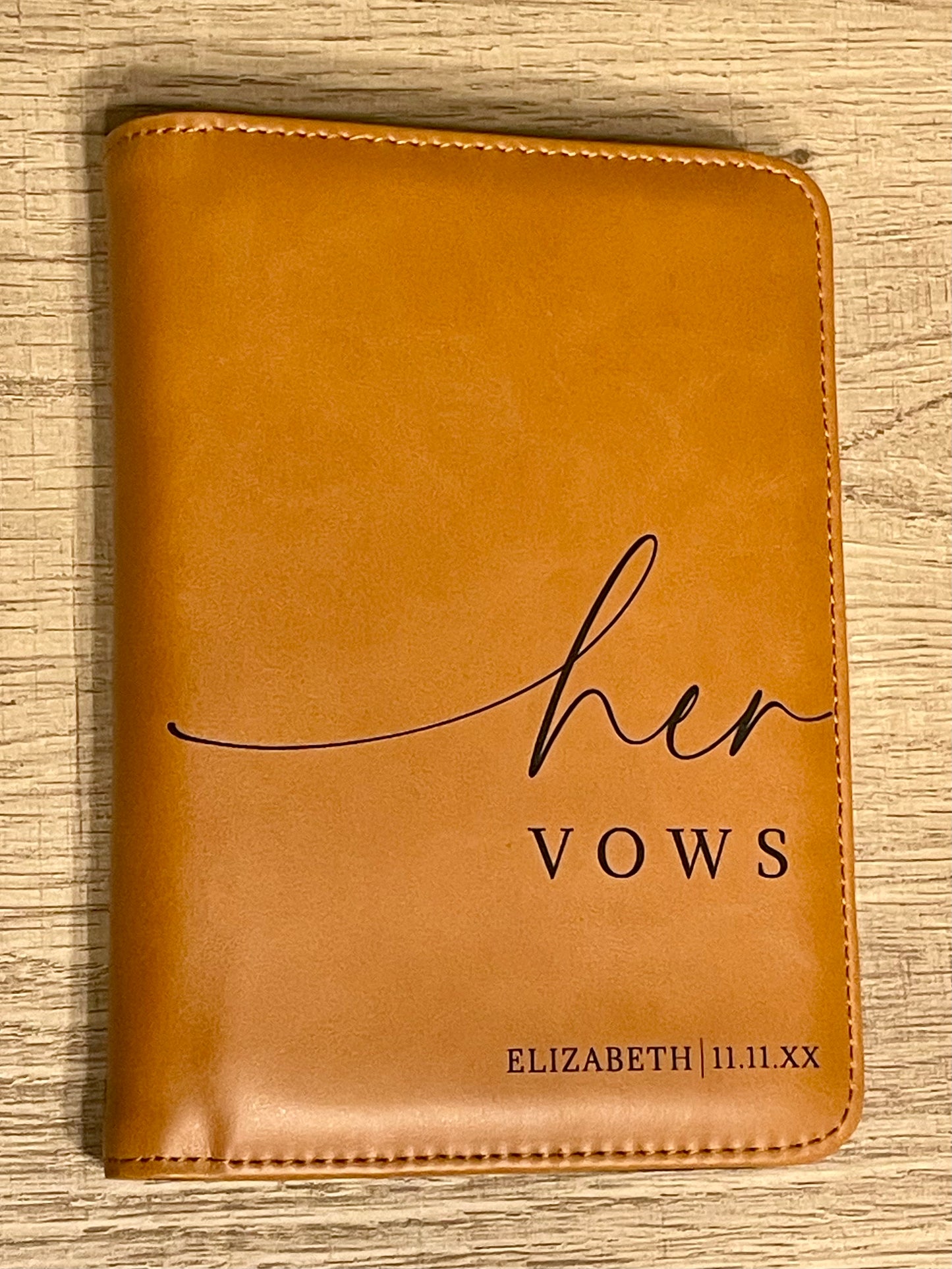 A rawhide vow book with "her VOWS ELIZABETH l 11.11.XX" engraved on the front, placed on a light wood surface.