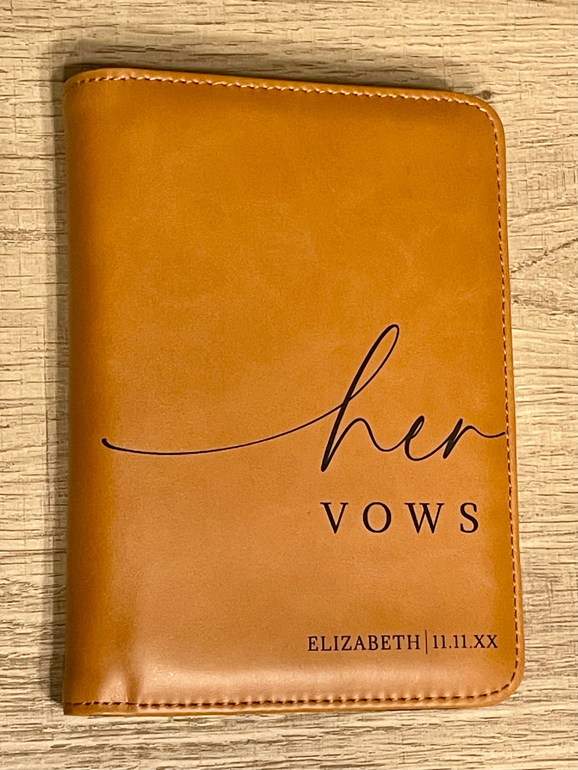 A rawhide vow book with "her VOWS ELIZABETH l 11.11.XX" engraved on the front, placed on a light wood surface.