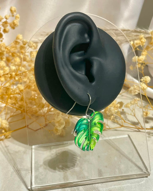 A pair of green monstera dangle earrings displayed on a black ear-shaped jewelry stand, set against a soft beige background with dried flowers.