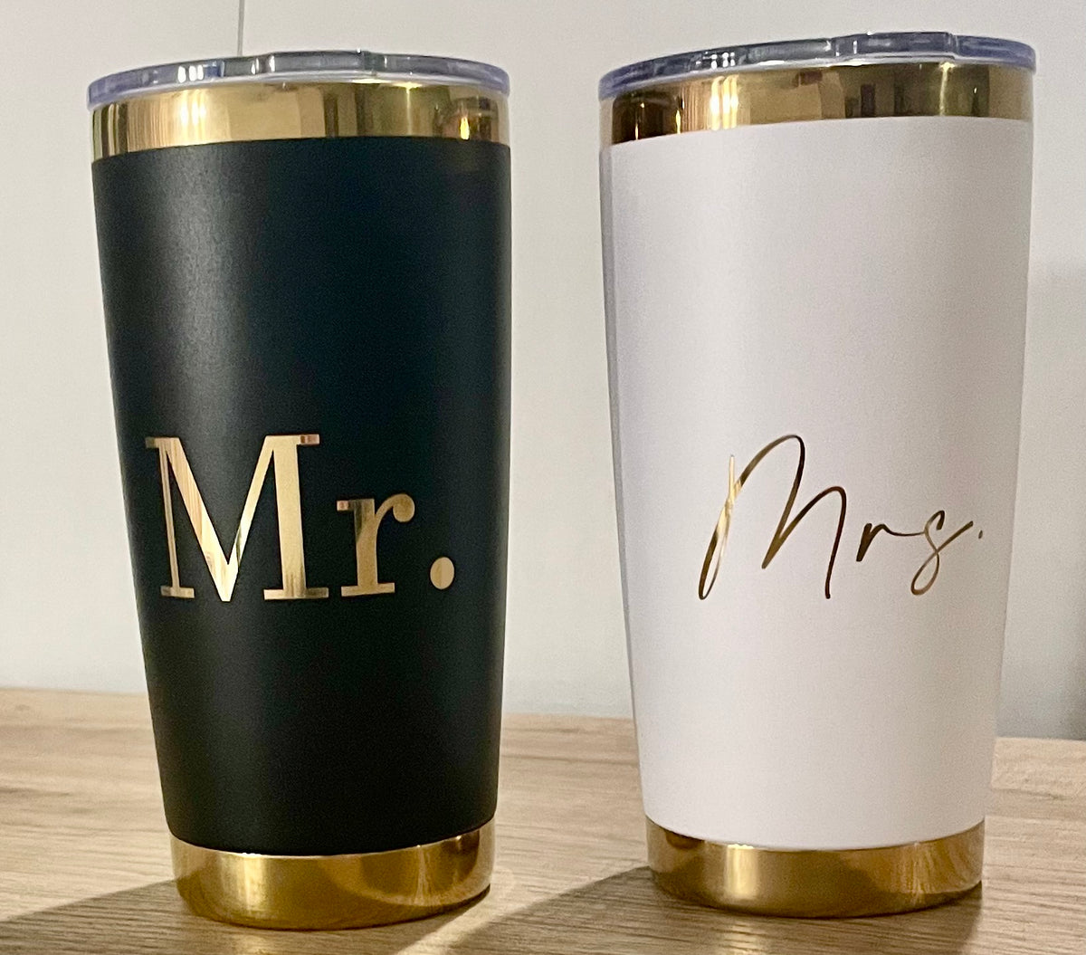 Two insulated stainless steel tumbler cups with clear lids on a wooden surface against a neutral wall. The left tumbler is matte black with metallic gold trim at the top and bottom and the word 'Mr.